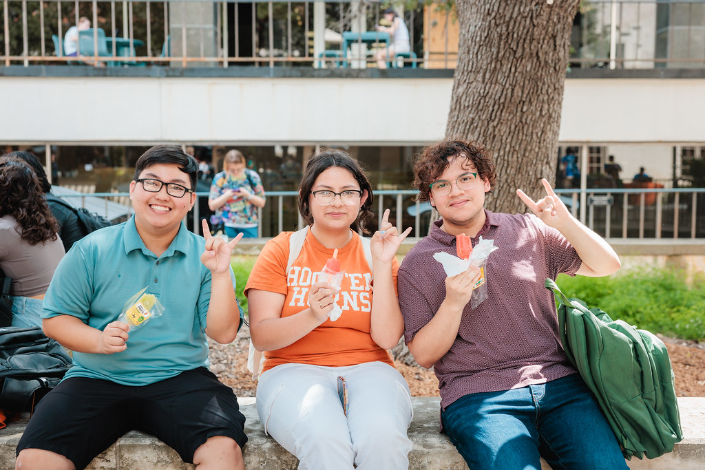  photo of three UT Austin students enjoying paletas on the Texas Union patio. The students are all smiling and flashing a hook 'em to the camera.